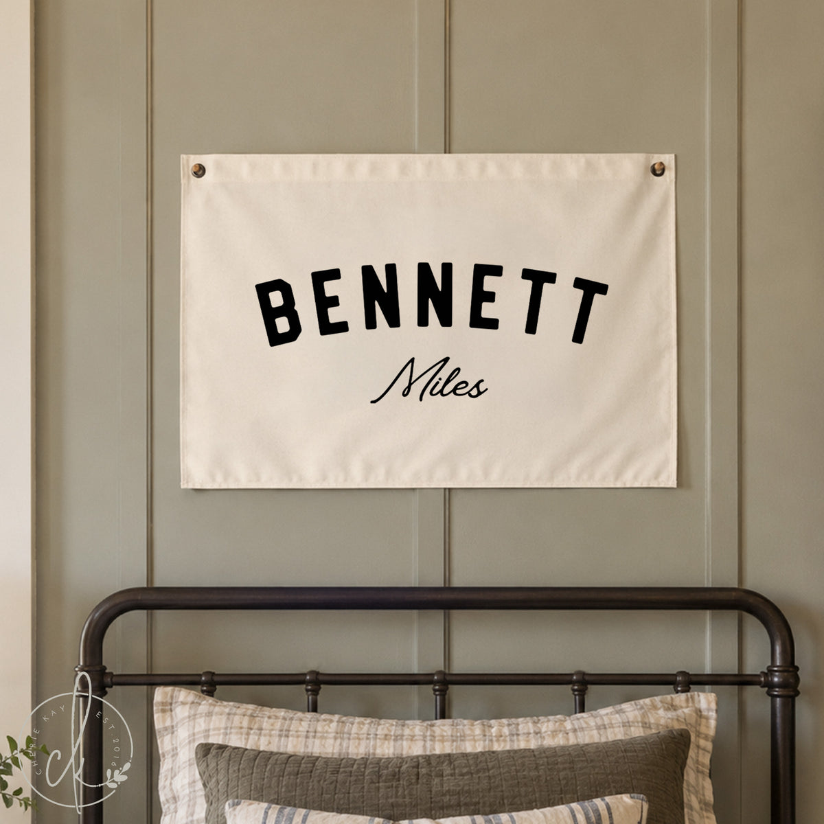 A personalized canvas name flag with the name Bennett Miles is displayed above a crib in a nursery room. The nursery features white walls, a crib with beige bedding, and a cozy blanket draped over the crib.