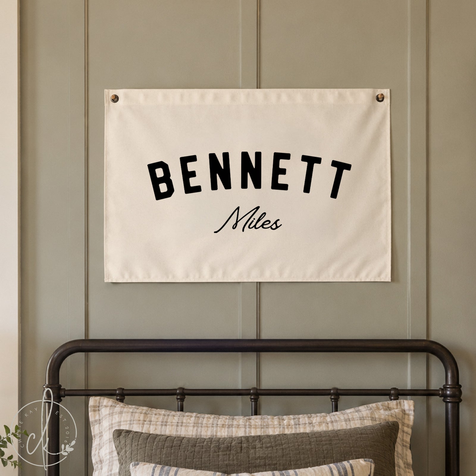 A personalized canvas name flag with the name Bennett Miles is displayed above a crib in a nursery room. The nursery features white walls, a crib with beige bedding, and a cozy blanket draped over the crib.