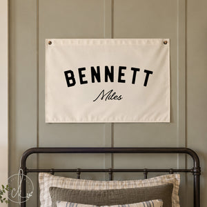 A personalized canvas name flag with the name Bennett Miles is displayed above a crib in a nursery room. The nursery features white walls, a crib with beige bedding, and a cozy blanket draped over the crib.
