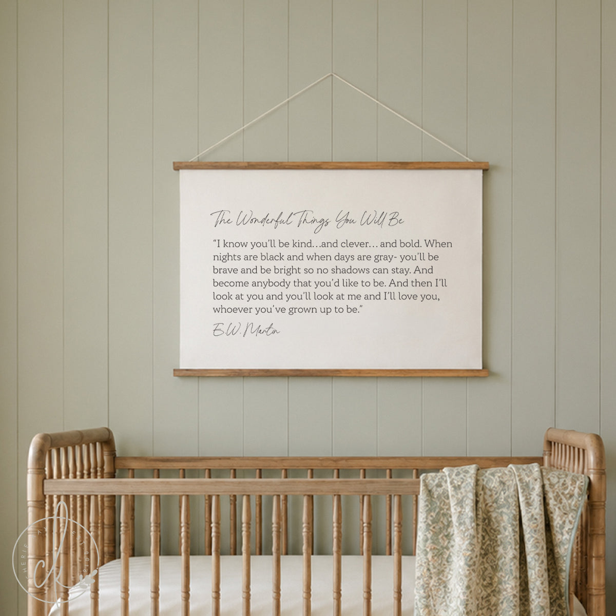 A cozy nursery features a white crib adorned with a floral pillow and a pink blanket draped over the side. Above the crib hangs a canvas flag with a quote by E.W. Martin printed in elegant script, adding an inspirational touch to the room's decor.