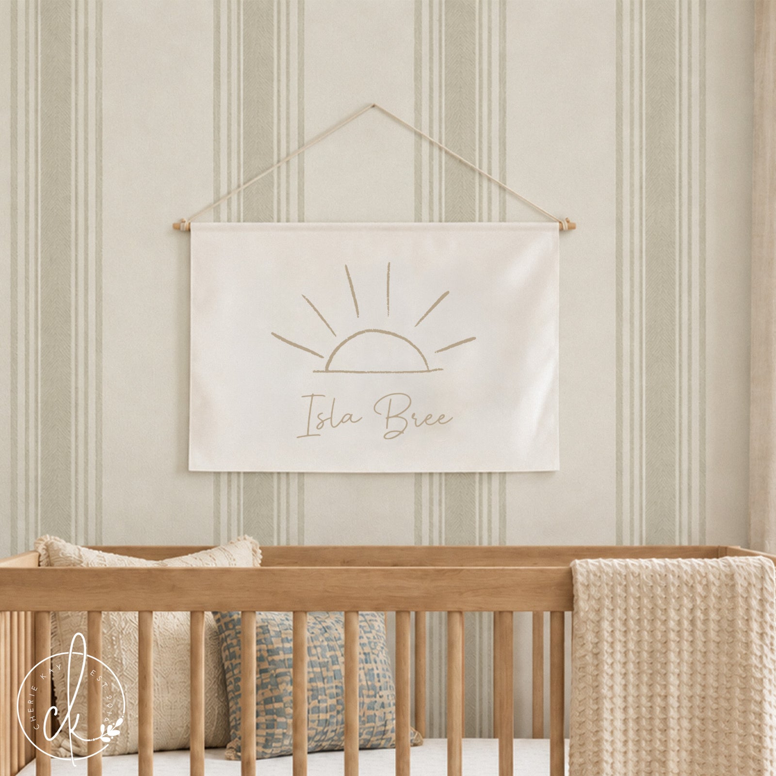 A stylish nursery featuring a light wooden crib with beige cushions and a cream blanket, set against a light green striped wall. Above the crib hangs a personalized canvas flag with a minimalist sun sketch and the name "Isla Bree" written below.