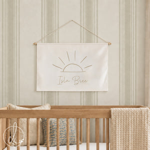 A stylish nursery featuring a light wooden crib with beige cushions and a cream blanket, set against a light green striped wall. Above the crib hangs a personalized canvas flag with a minimalist sun sketch and the name "Isla Bree" written below.