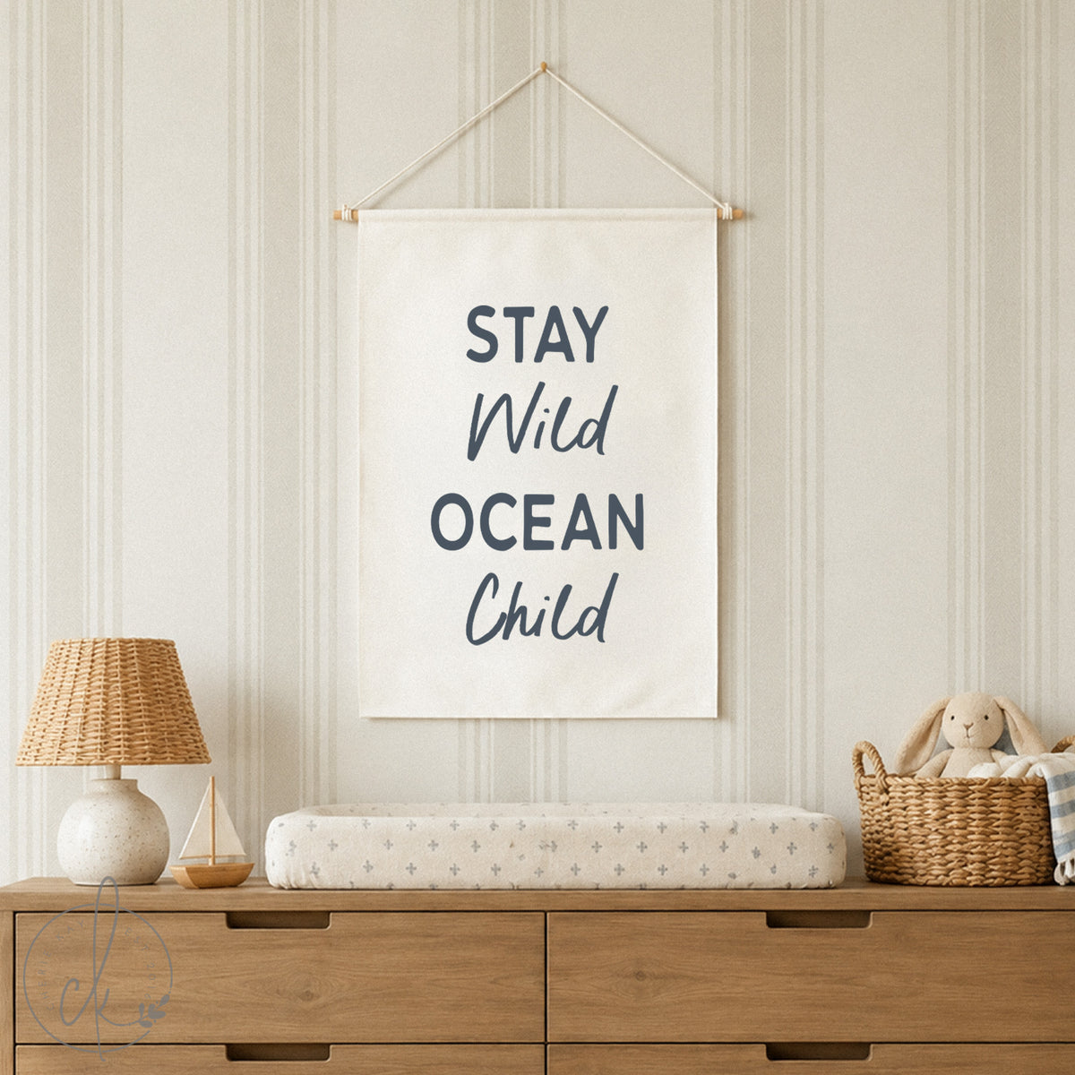 A nursery corner with a white crib and beige blanket, a surfboard, and a potted plant against a green wall. Above the crib hangs a canvas sign reading "Stay Wild Ocean Child."