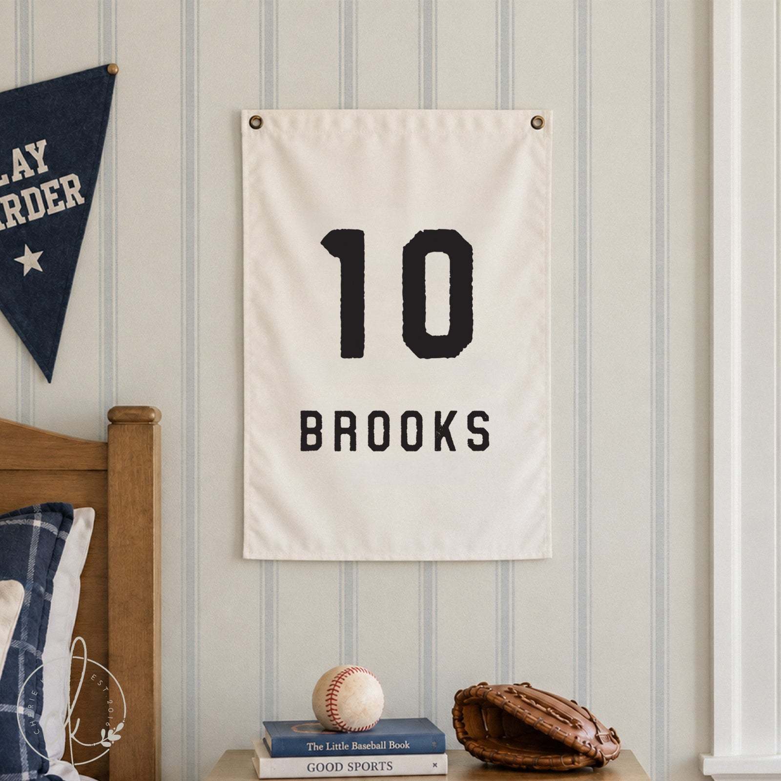 A personalized sports-themed sign featuring a large 10 and the name BROOKS displayed in bold, black typography on a neutral hanging sign. The sign is hung  on a striped wall above a boys nightstand. 