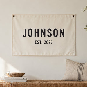 Personalized family name canvas sign, displayed on a rustic brick wall with greenery. Custom established date wall decor, perfect for anniversary, wedding, housewarming, or family gift.