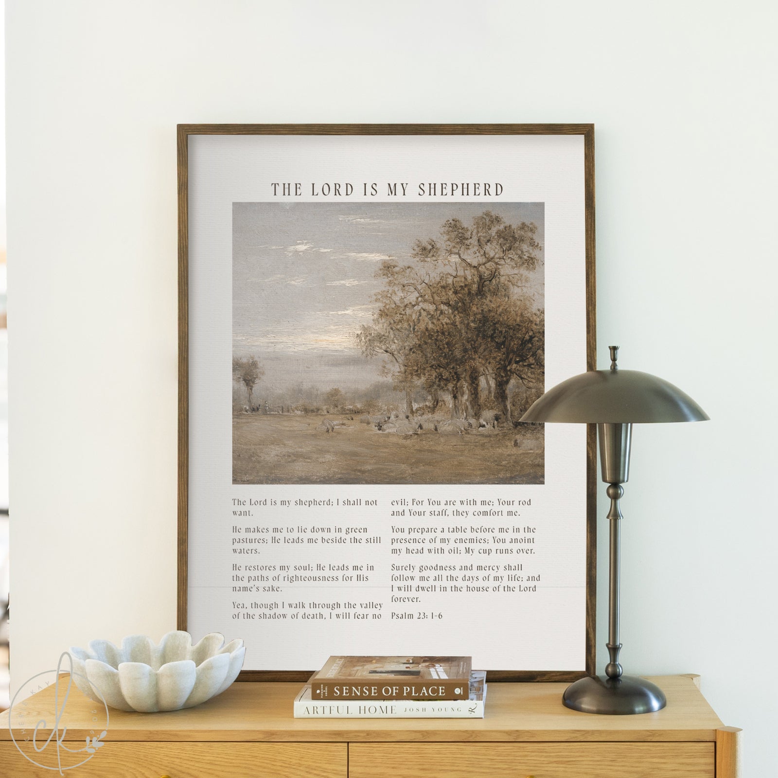 Framed print with pastoral scene and text on a wooden cabinet against a white wall.