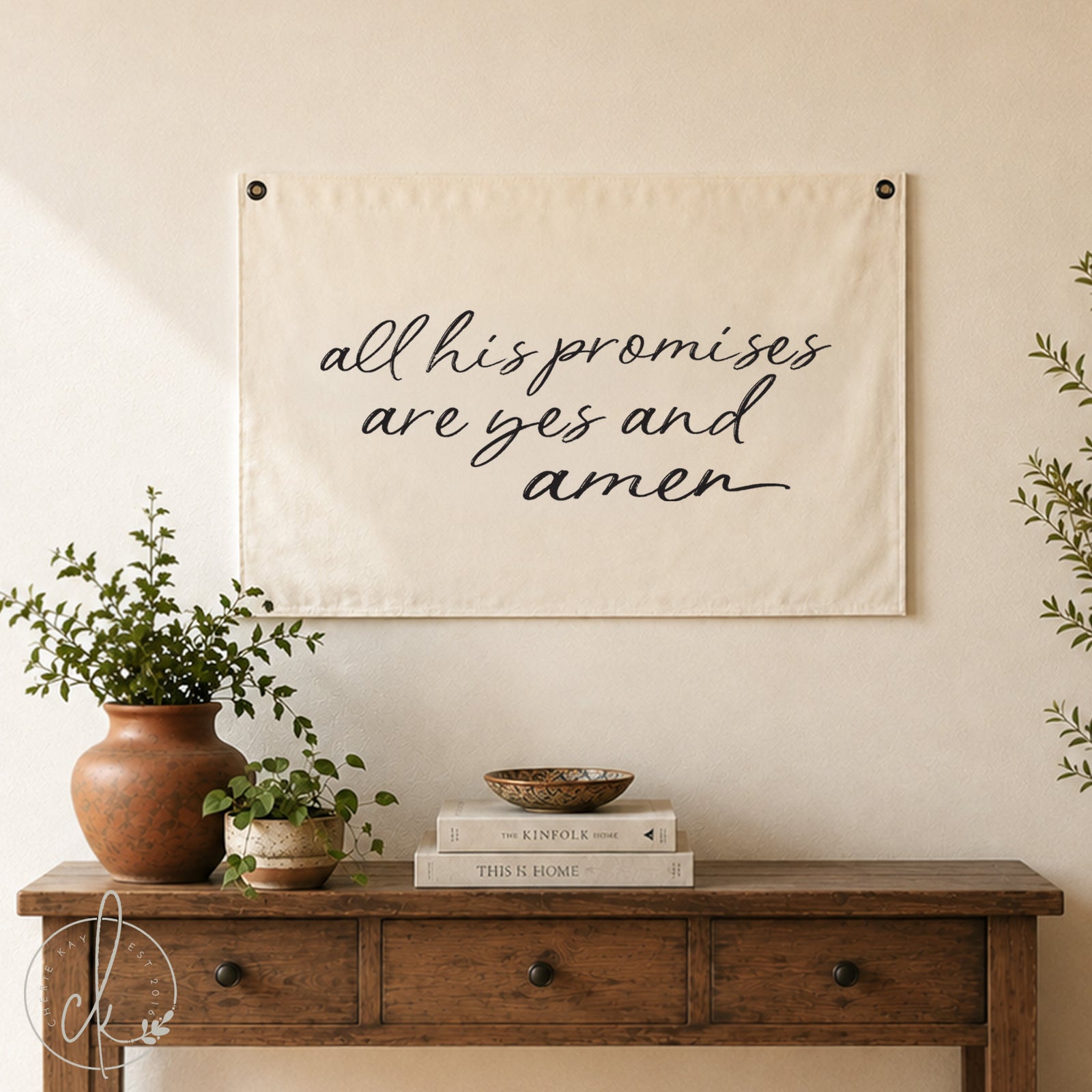 Decorative wooden dresser with a potted plant, vintage clock, and books, with a fabric wall banner hanging above it that reads All His Promises Are Yes and Amen in elegant script.