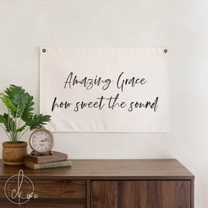 Decorative banner with 'Amazing Grace, how sweet the sound' text above a wooden desk with a plant and clock.