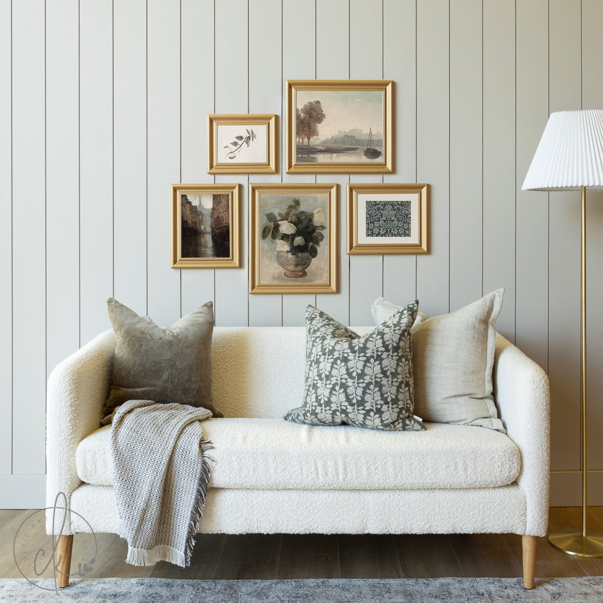 Curated vintage gallery wall featuring framed botanical and landscape art prints in gold frames, styled above a cozy cream sofa with decorative pillows in a living room setting.