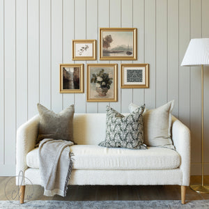 Curated vintage gallery wall featuring framed botanical and landscape art prints in gold frames, styled above a cozy cream sofa with decorative pillows in a living room setting.