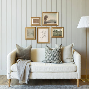 Cozy living room with a gold-framed gallery wall art set featuring minimalist botanical and landscape prints, arranged above a cream-colored textured sofa with decorative pillows and a throw blanket, creating a warm and elegant ambiance.
