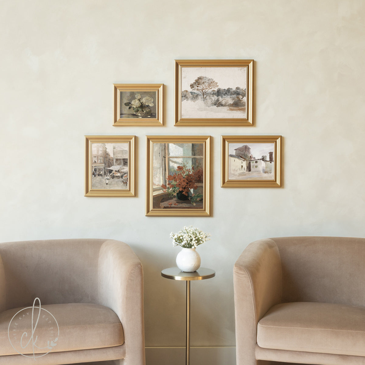 A cozy seating area features two beige upholstered chairs flanking a small gold side table with white flowers, beneath a gallery wall of five gold-framed vintage paintings and prints. The artwork includes floral still lifes, landscapes, and architectural scenes arranged in an asymmetrical pattern against a cream-colored wall.