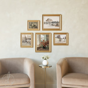 A cozy seating area features two beige upholstered chairs flanking a small gold side table with white flowers, beneath a gallery wall of five gold-framed vintage paintings and prints. The artwork includes floral still lifes, landscapes, and architectural scenes arranged in an asymmetrical pattern against a cream-colored wall.