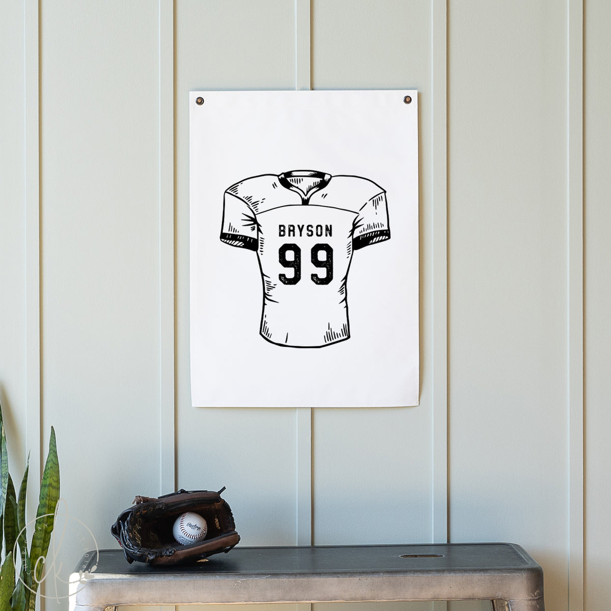 White wall with a framed black and white illustration of a football jersey with the name &#39;Grayson&#39; and number &#39;99&#39;. Below, there is a baseball glove and ball on a wooden surface.