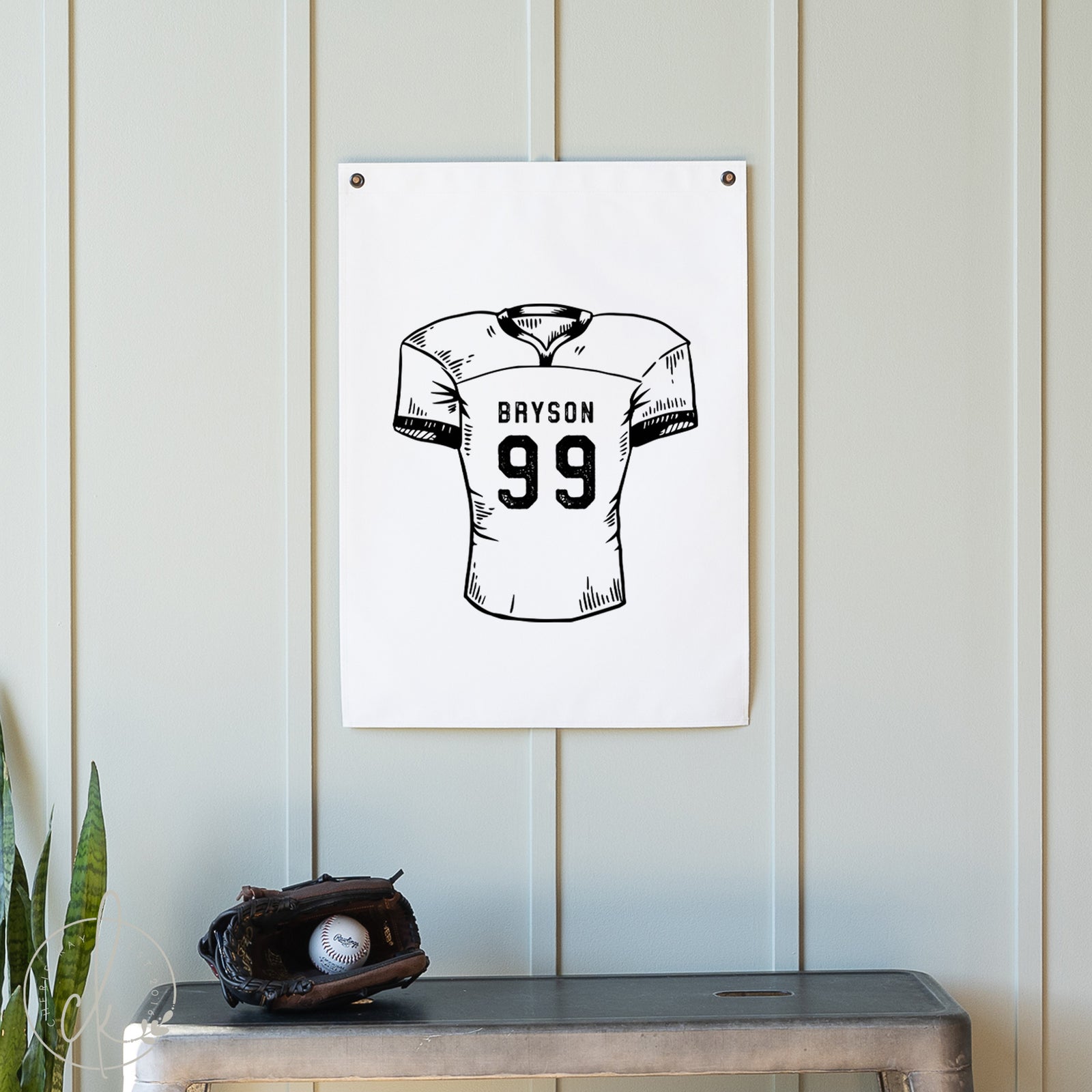 White wall with a framed black and white illustration of a football jersey with the name 'Grayson' and number '99'. Below, there is a baseball glove and ball on a wooden surface.