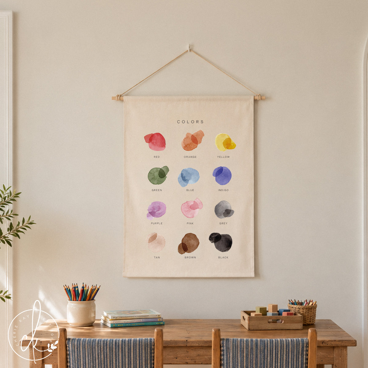 A rectangular fabric wall hanging featuring the word COLORS at the top, followed by twelve watercolor-style paint splatters in red, orange, yellow, green, blue, indigo, purple, pink, grey, tan, brown, and black, each labeled below.