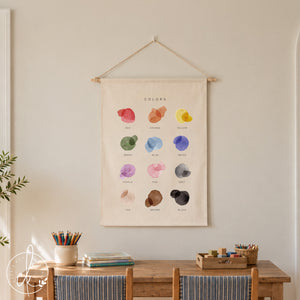 A rectangular fabric wall hanging featuring the word COLORS at the top, followed by twelve watercolor-style paint splatters in red, orange, yellow, green, blue, indigo, purple, pink, grey, tan, brown, and black, each labeled below.