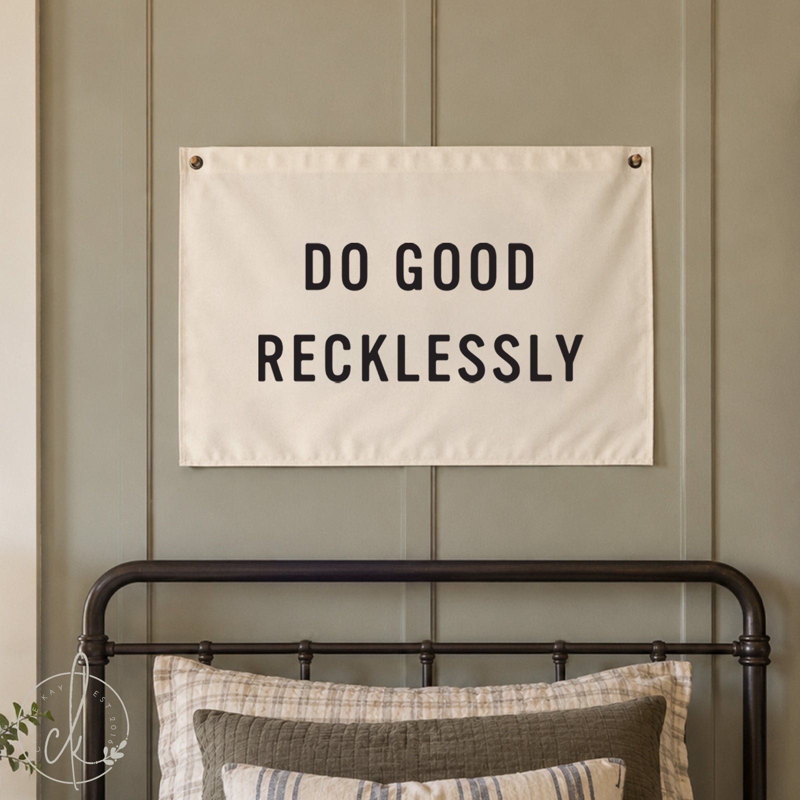 A cream canvas wall banner with the black text 'DO GOOD RECKLESSY' hanging on a muted sage green paneled wall above a black metal bed frame with pillows.
