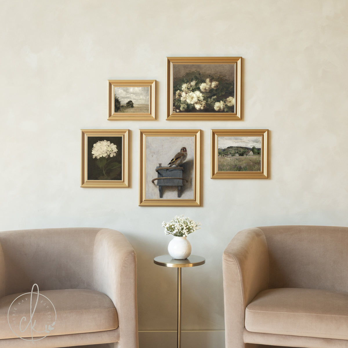 Five gold-framed vintage artworks displayed in an asymmetrical gallery wall arrangement above beige velvet armchairs, featuring a mix of floral still lifes, countryside landscapes, and a charming bird perched on books, creating an elegant focal point in a neutral-toned living space.