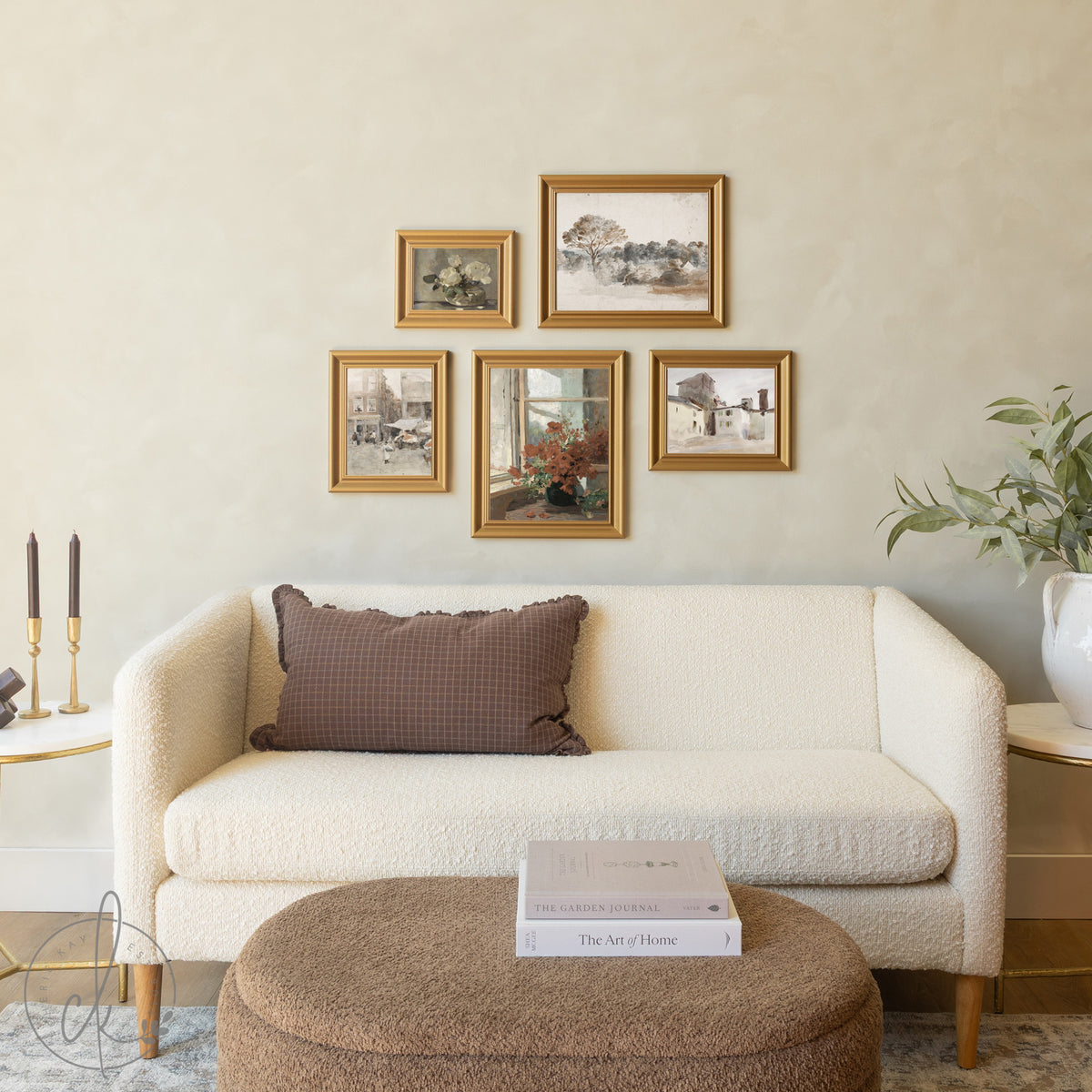 A cream-colored modern sofa with a brown checkered throw pillow sits beneath a gallery wall of five gold-framed vintage artworks, with a round upholstered ottoman holding design books in front and decorative elements like candles and a potted plant nearby.