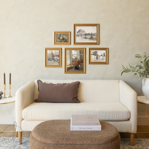 A cream-colored modern sofa with a brown checkered throw pillow sits beneath a gallery wall of five gold-framed vintage artworks, with a round upholstered ottoman holding design books in front and decorative elements like candles and a potted plant nearby.