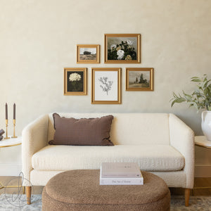 A collection of five gold-framed botanical and landscape artworks arranged in a gallery wall style above a cream textured sofa, featuring floral paintings, a botanical print, and pastoral scenes, with a round upholstered ottoman and decorative accessories completing the cozy living space.RetryClaude can make mistakes. Please double-check responses.