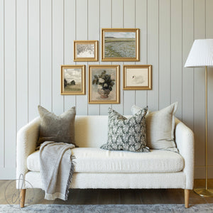 Cozy living room with a rustic gallery wall featuring five gold-framed art prints, including vintage landscapes, botanicals, and classic artwork, above a neutral sofa with accent pillows and a throw blanket.