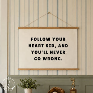 A white rectangular wall banner featuring the black text FOLLOW YOUR HEART KID, AND YOU'LL NEVER GO WRONG. The banner is mounted wood strips and string, and hangs over a boys dresser
