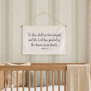 A rectangular off-white fabric wall hanging featuring the black printed text: For this child we have prayed and the Lord has granted us the desires of our hearts 1 SAMUEL 1:27. The banner is suspended by a thin white string.