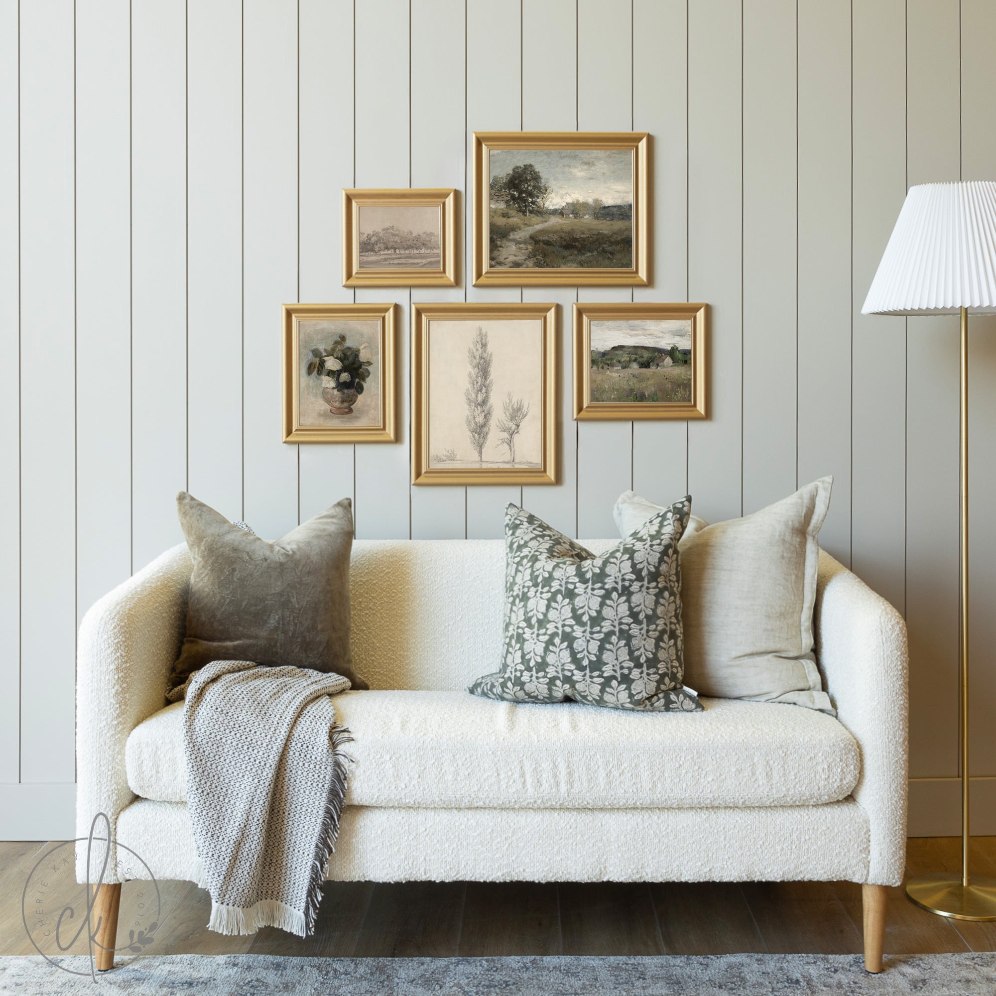 A cozy living room setup featuring a modern farmhouse-style gallery wall with five framed nature-inspired art prints, a cream textured sofa adorned with neutral and floral-patterned pillows, a soft throw blanket, and a standing lamp in the corner.