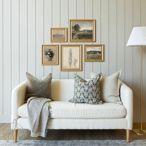 A cozy living room setup featuring a modern farmhouse-style gallery wall with five framed nature-inspired art prints, a cream textured sofa adorned with neutral and floral-patterned pillows, a soft throw blanket, and a standing lamp in the corner.