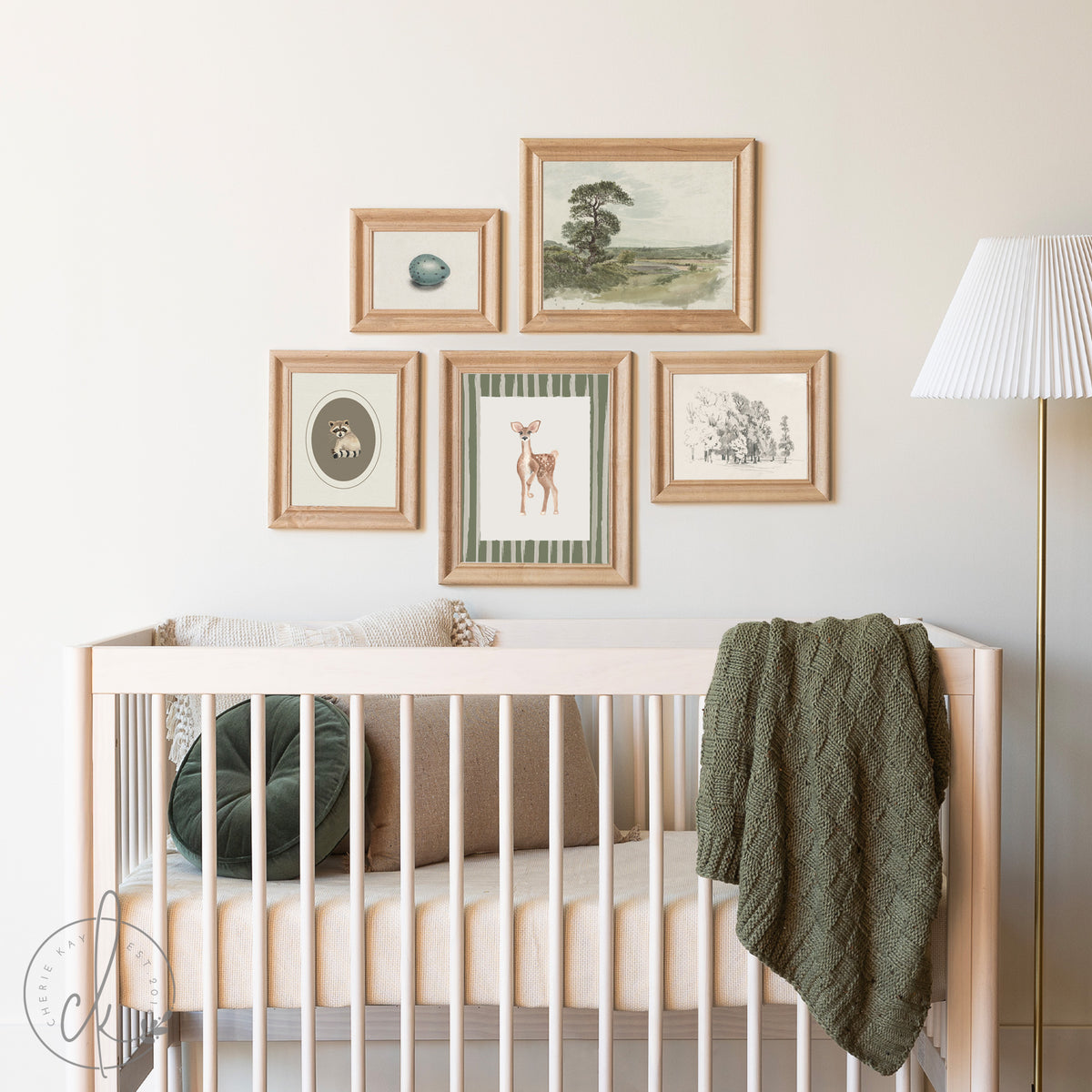 Rustic woodland nursery featuring a crib with neutral bedding, a green knitted blanket, and a gallery wall of framed art prints, including a fawn, raccoon, bird egg, and nature landscapes, in natural wood frames.