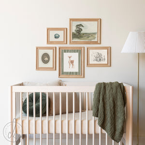 Rustic woodland nursery featuring a crib with neutral bedding, a green knitted blanket, and a gallery wall of framed art prints, including a fawn, raccoon, bird egg, and nature landscapes, in natural wood frames.