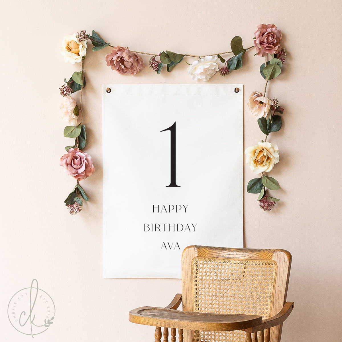 Floral garland with a &#39;Happy Birthday Ava&#39; sign on a beige wall with a wooden chair.