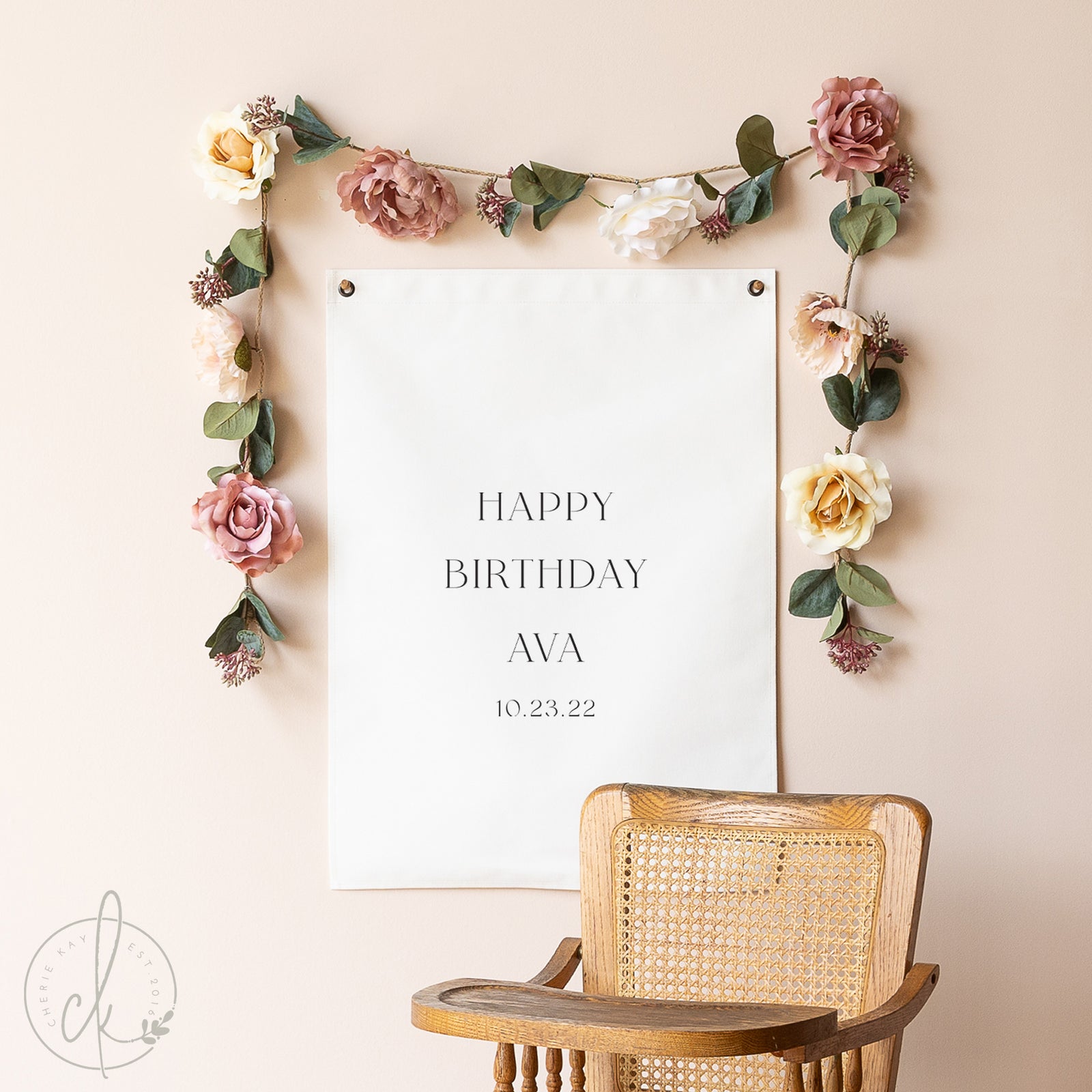 Floral garland with a 'Happy Birthday Ava' sign on a beige wall with a wooden chair.