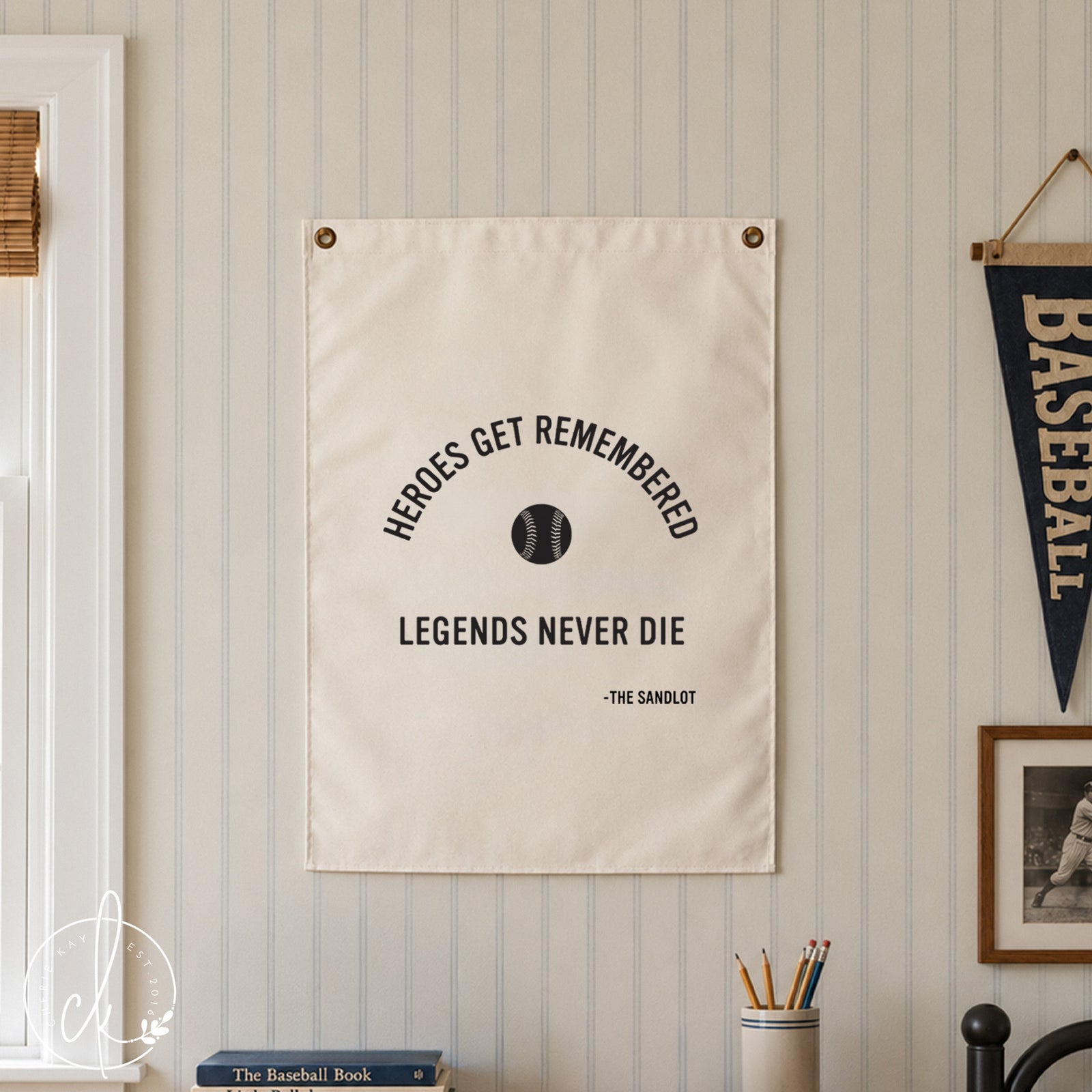 A white rectangular wall hanging featuring the black text HEROES GET REMEMBERED, a small black baseball graphic, the text LEGENDS NEVER DIE, and the attribution -THE SANDLOT, secured with two metal grommets at the top corners.