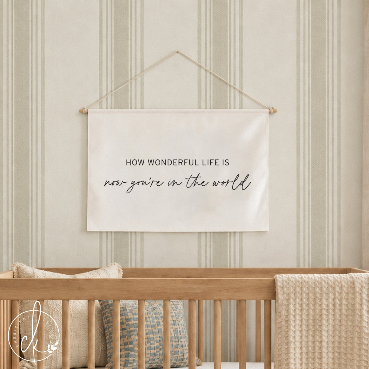 A rectangular off-white fabric wall hanging with a string for suspension. The text reads: HOW WONDERFUL LIFE IS now you're in the world. It is positioned above a black crib containing two beige pillows and a textured beige throw blanket.