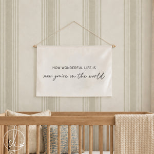 A rectangular off-white fabric wall hanging with a string for suspension. The text reads: HOW WONDERFUL LIFE IS now you're in the world. It is positioned above a black crib containing two beige pillows and a textured beige throw blanket.