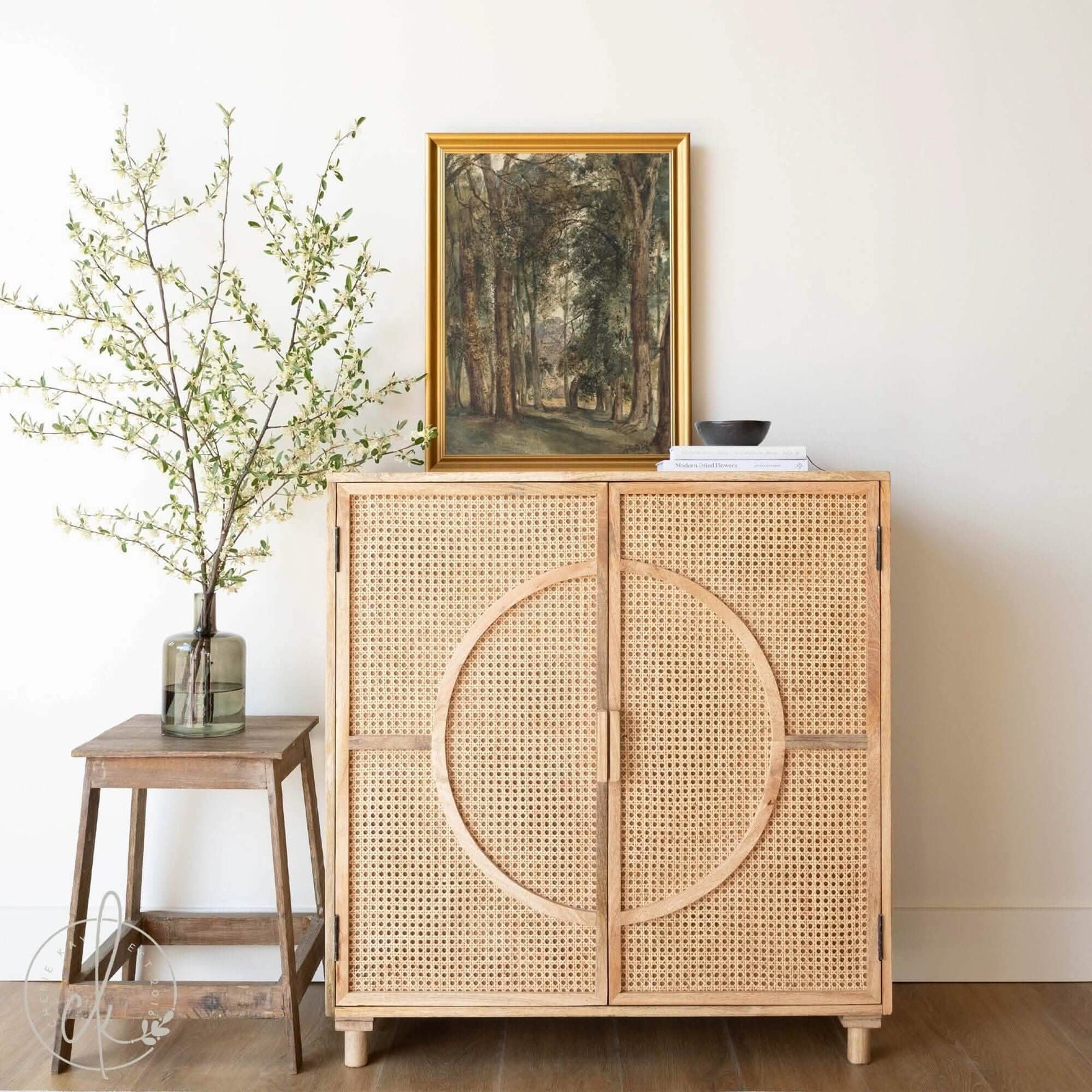 A beautifully styled living space featuring a vintage forest path landscape painting in a gold frame displayed on top of a natural rattan cabinet. Adding a touch of nature and elegance to the room decor. A blend of vintage and modern rustic elements.