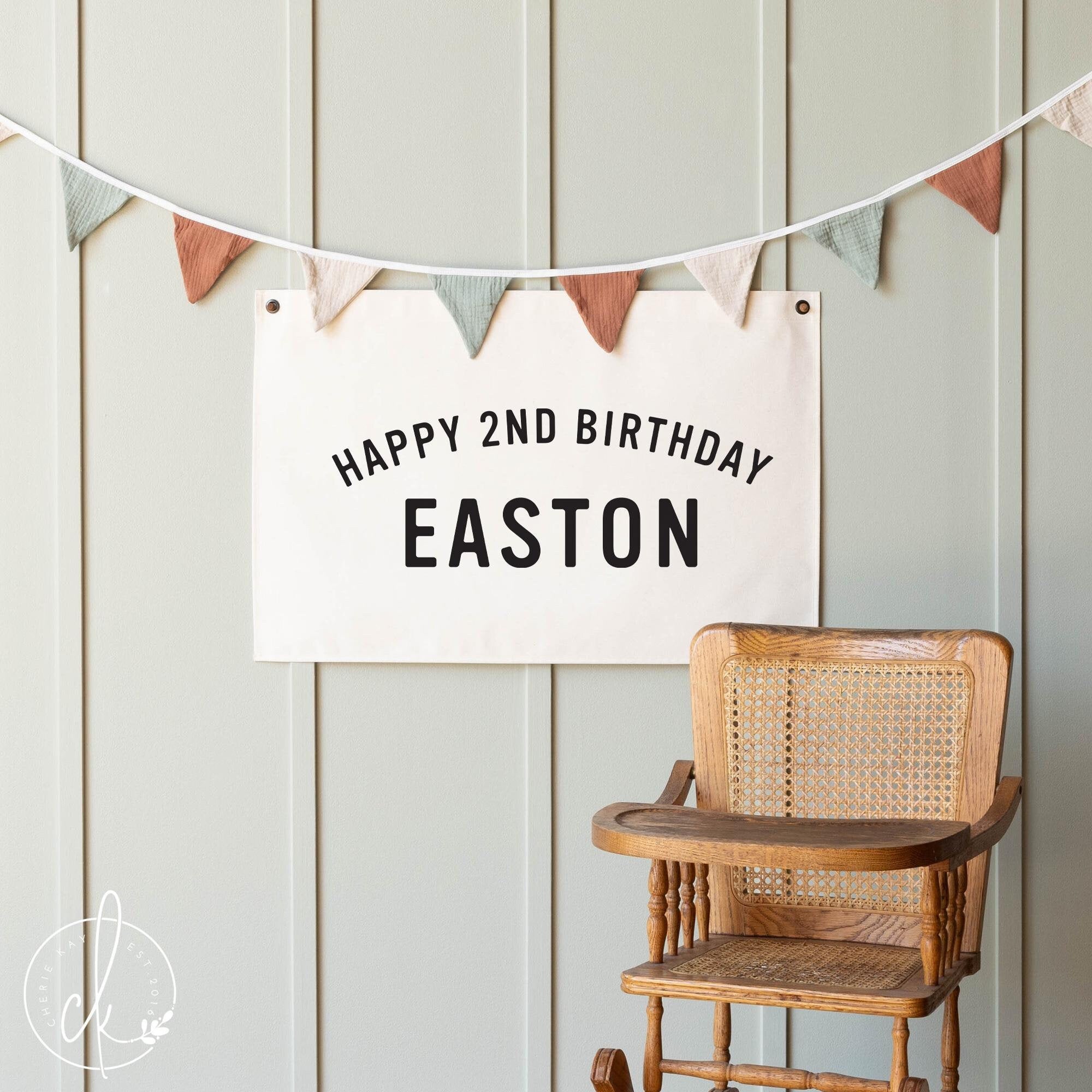 Custom Happy Birthday banner with personalized name in bold black text on a neutral canvas backdrop, displayed with colorful bunting above, ideal for toddler birthday party decor or photo shoots.