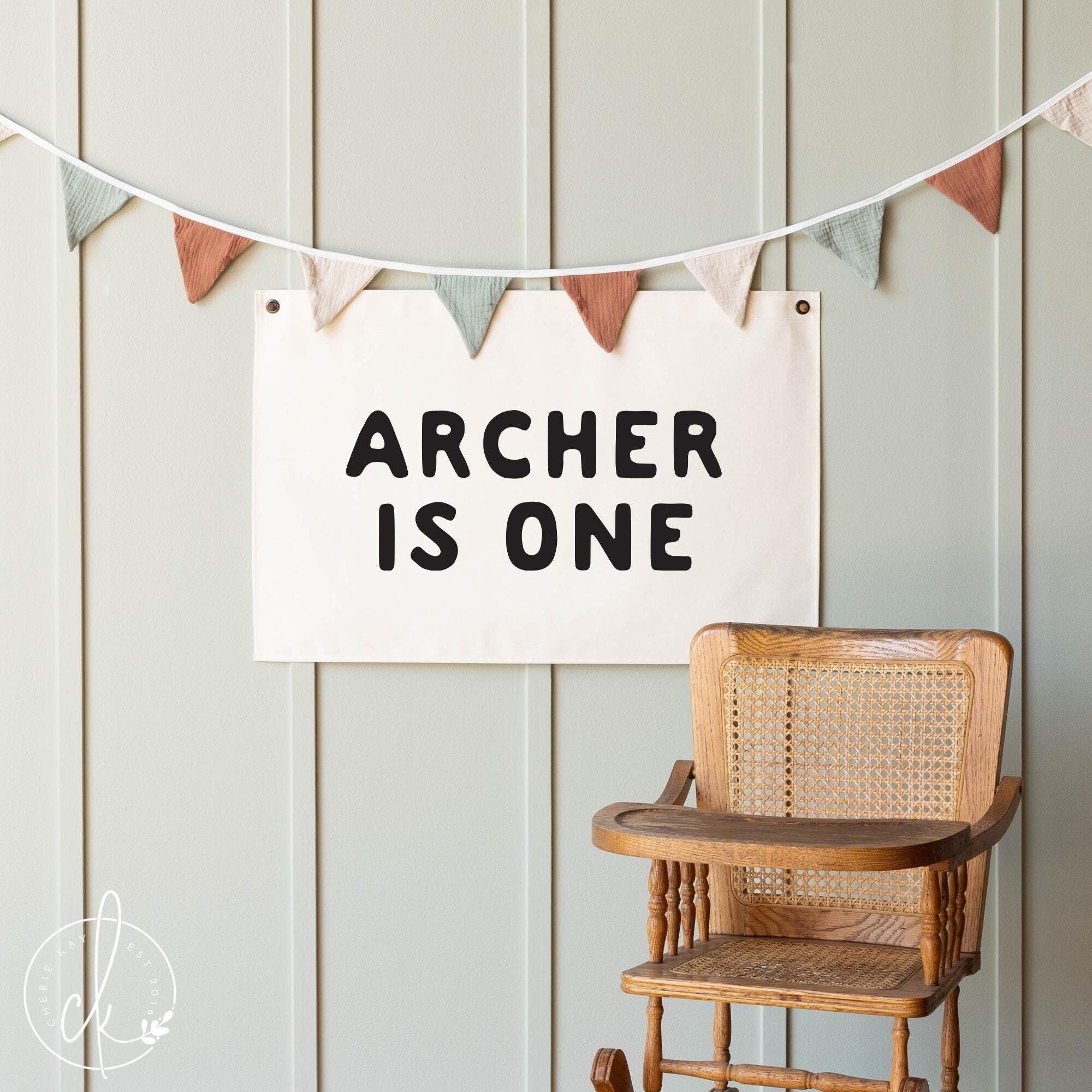 A personalized canvas birthday banner with the text Archer is One is displayed on a light-colored wall. Ideal custom decor for a child&#39;s first birthday party, or milestone celebration.