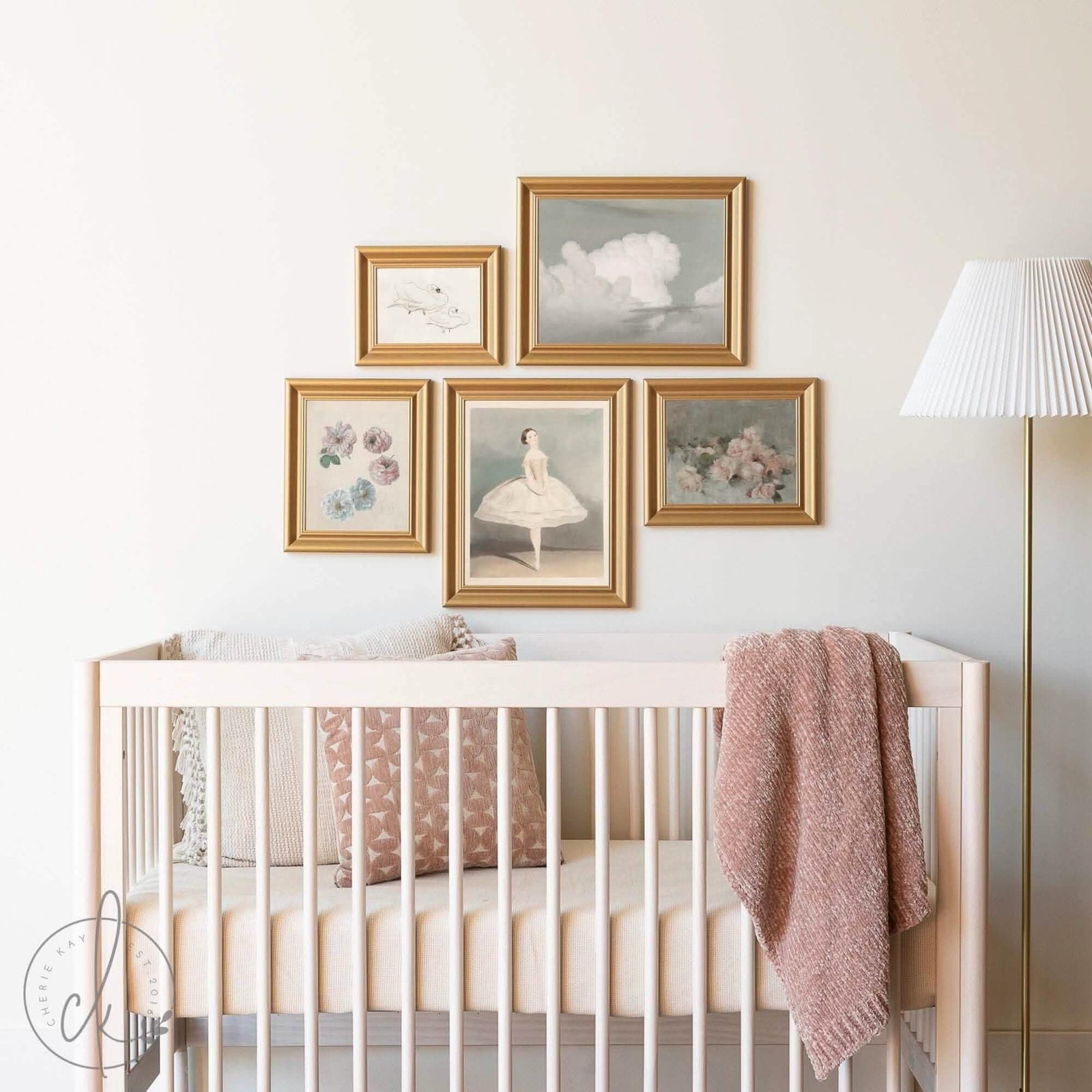 A nursery with a light wooden crib, soft bedding, and a pink blanket. Above the crib, a gallery wall features five gold-framed art prints, including florals, a ballet dancer, and clouds. A white floor lamp stands nearby.
