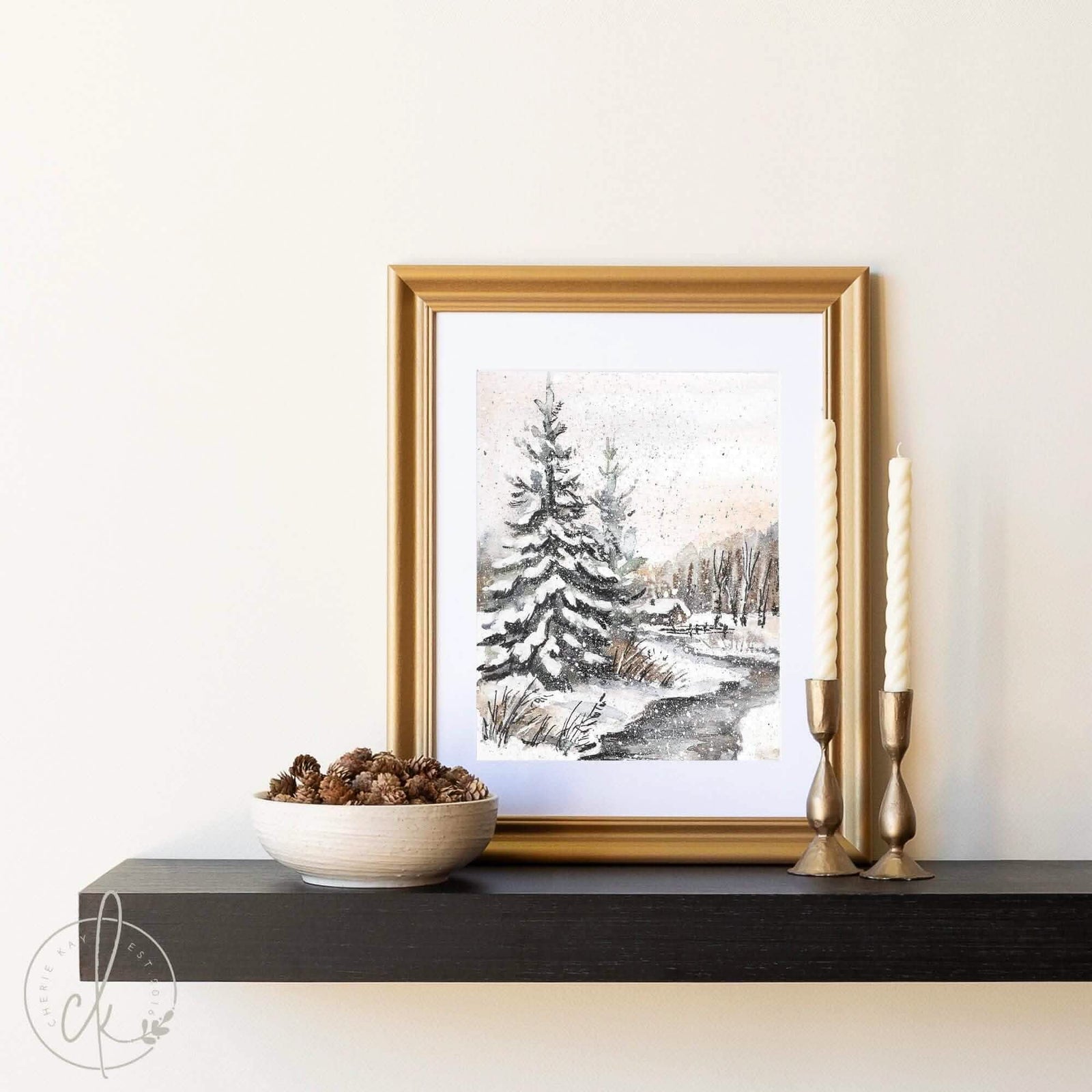 Framed winter landscape art print with a snowy pine tree scene, displayed on a black shelf with gold candle holders and a bowl of pinecones, creating a cozy holiday decor setting.