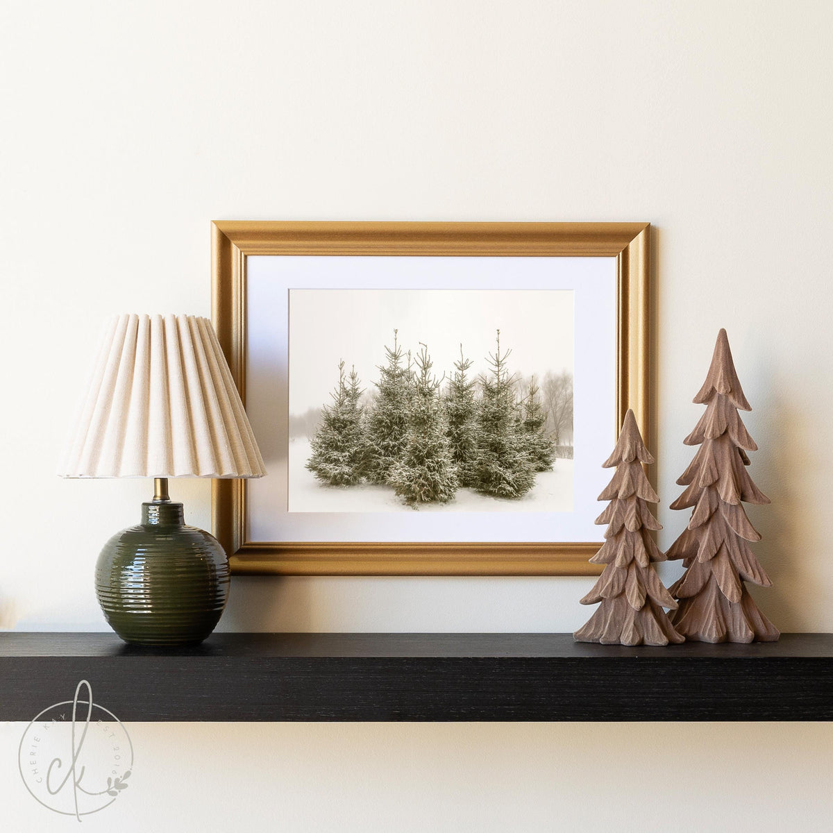 Framed winter landscape art print featuring a serene forest scene with snow-covered trees, displayed on a black shelf alongside a table lamp and decorative wooden trees, creating a cozy and rustic home decor setup