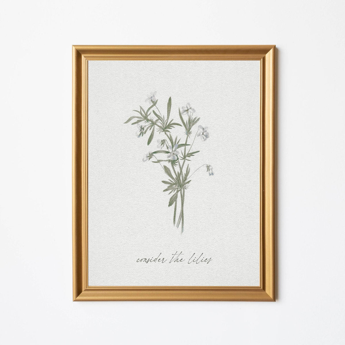 Framed artwork featuring a delicate botanical illustration of lilies with the phrase - consider the lilies - from Matthew 6:27 below, set in an elegant gold frame against a light-colored background.