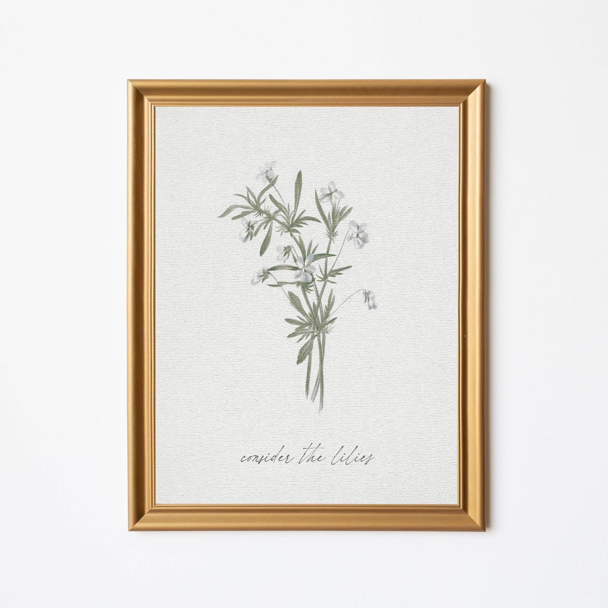 Framed artwork featuring a delicate botanical illustration of lilies with the phrase - consider the lilies - from Matthew 6:27 below, set in an elegant gold frame against a light-colored background.