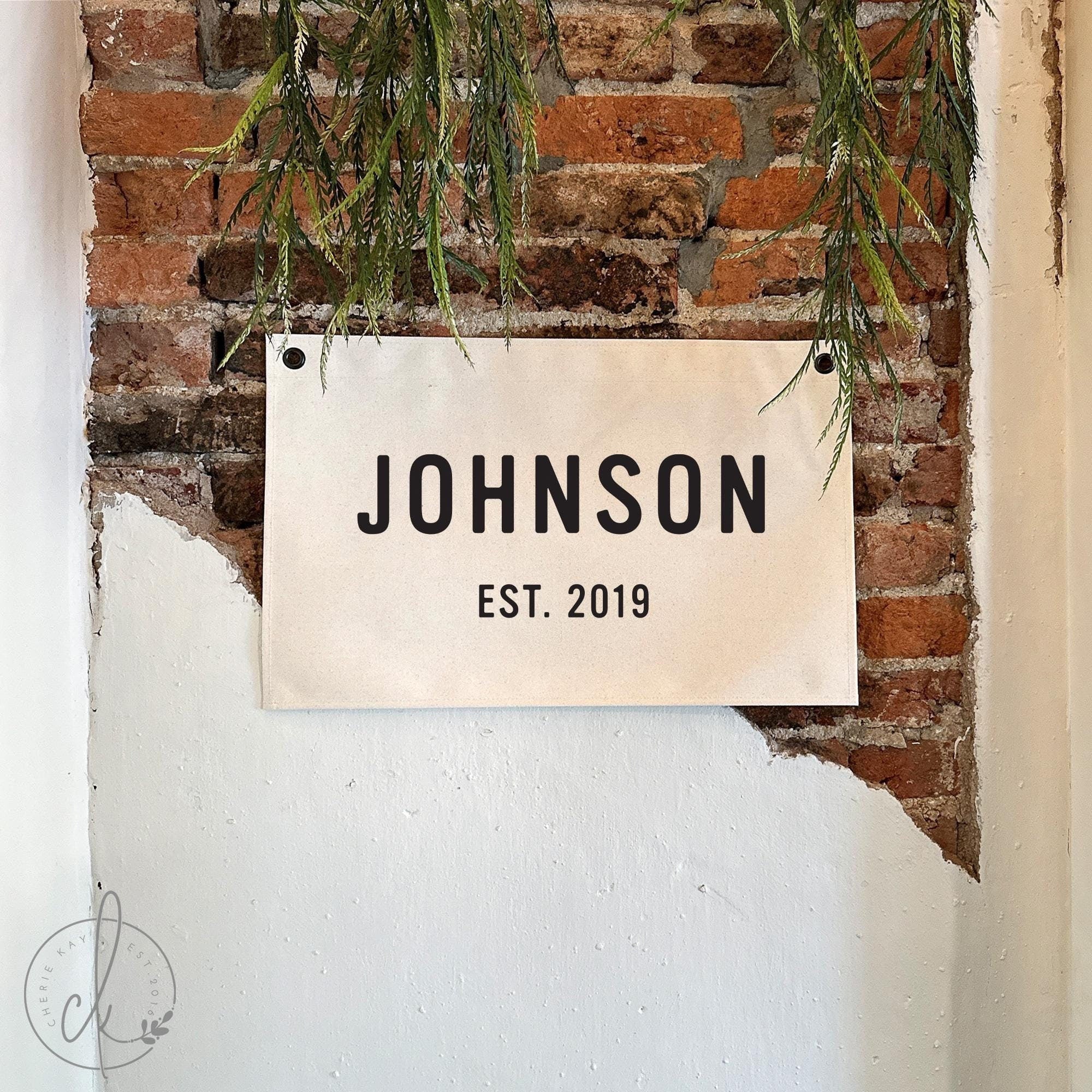 Personalized family name canvas sign, displayed on a rustic brick wall with greenery. Custom established date wall decor, perfect for anniversary, wedding, housewarming, or family gift.