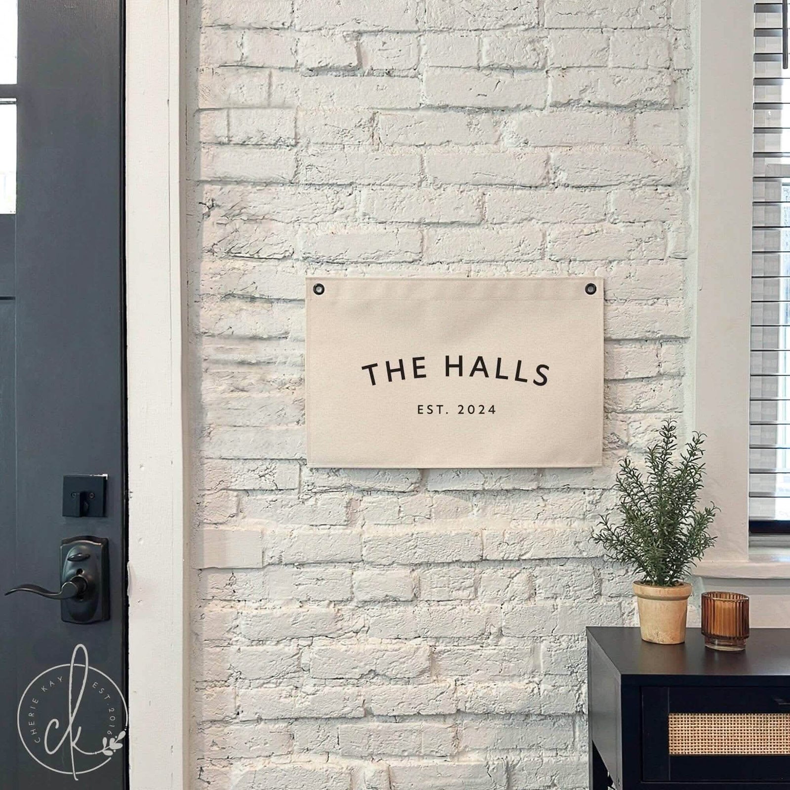 Personalized canvas banner hanging on a white brick wall, featuring the text &#39;The Halls, Est. 2024.&#39; The sign is displayed near a black side table with a small potted plant and candle, adding a modern rustic touch to the entryway decor.