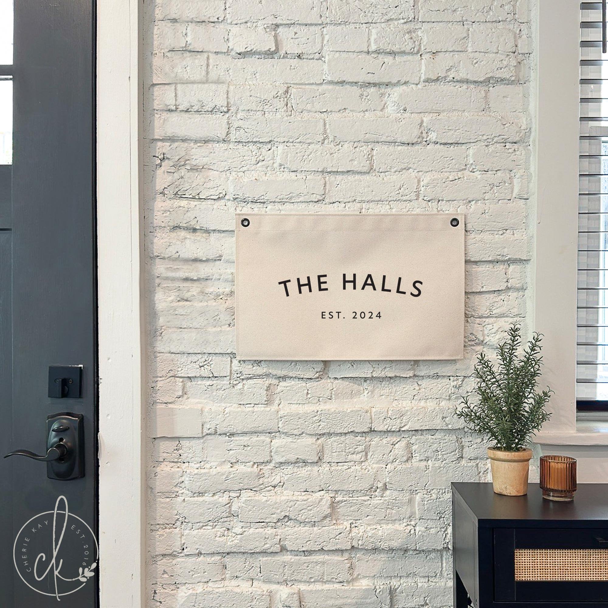 Personalized canvas banner hanging on a white brick wall, featuring the text &#39;The Halls, Est. 2024.&#39; The sign is displayed near a black side table with a small potted plant and candle, adding a modern rustic touch to the entryway decor.