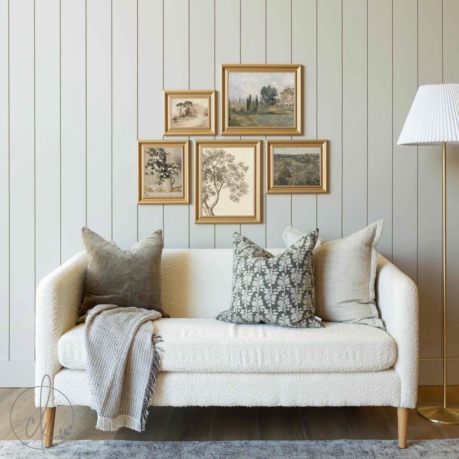 Cozy living room featuring a white textured sofa with decorative pillows and a throw blanket, accented by a gold-framed gallery wall set of vintage-inspired artwork, including landscapes, botanicals, and still-life pieces, against a shiplap wall.